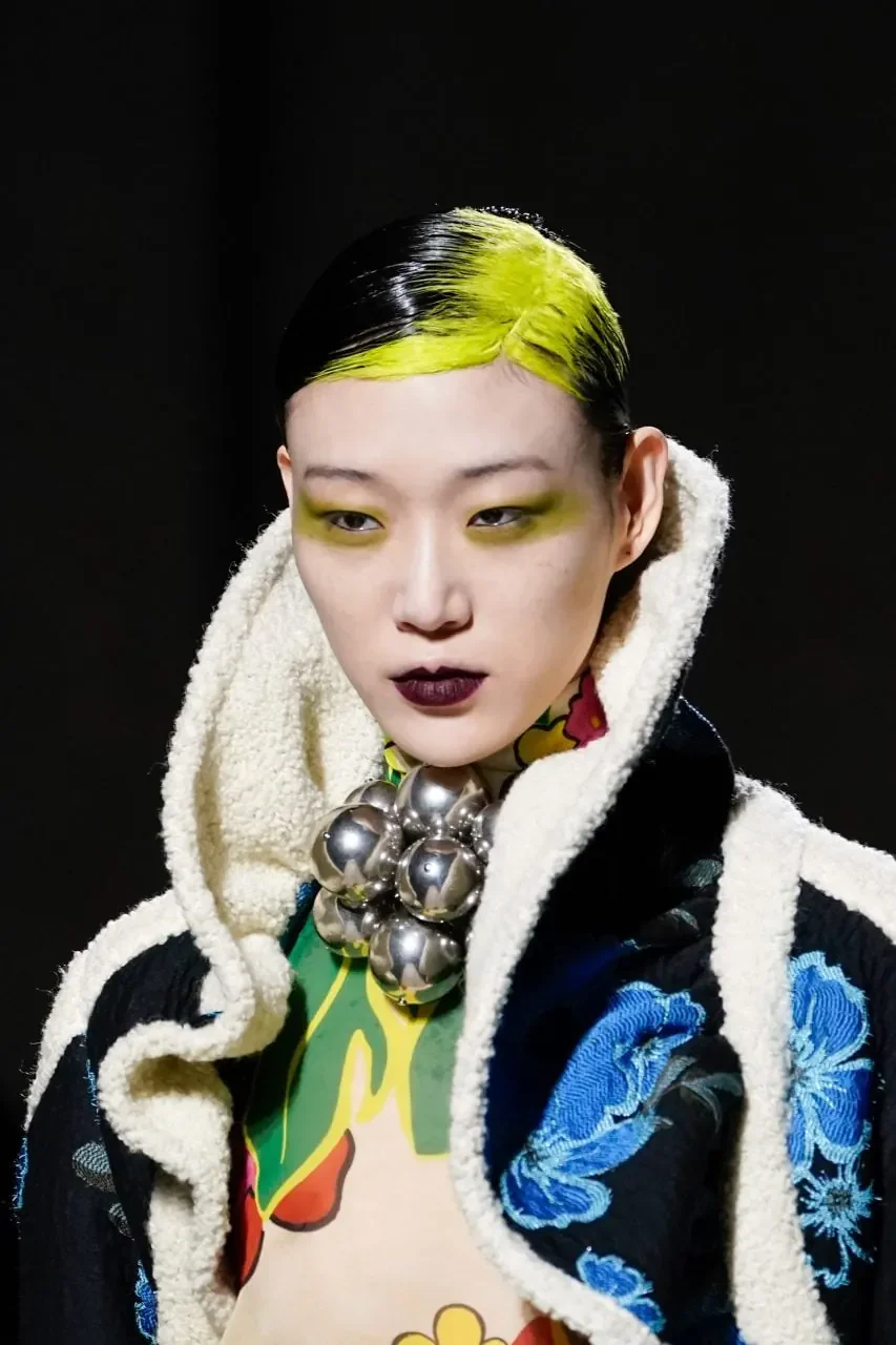 Woman with short slicked-back black hair and a broad neon-yellow section across the front hairline and crown roots.