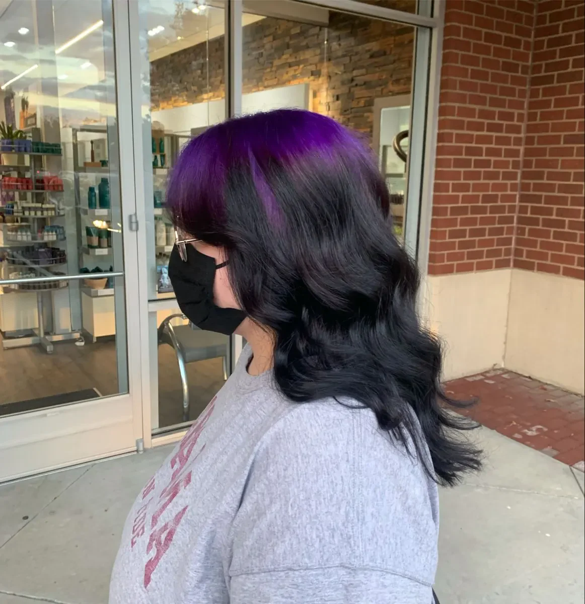 Side view of a woman with medium black body-wave hair and a violet-purple panel sweeping from the roots.