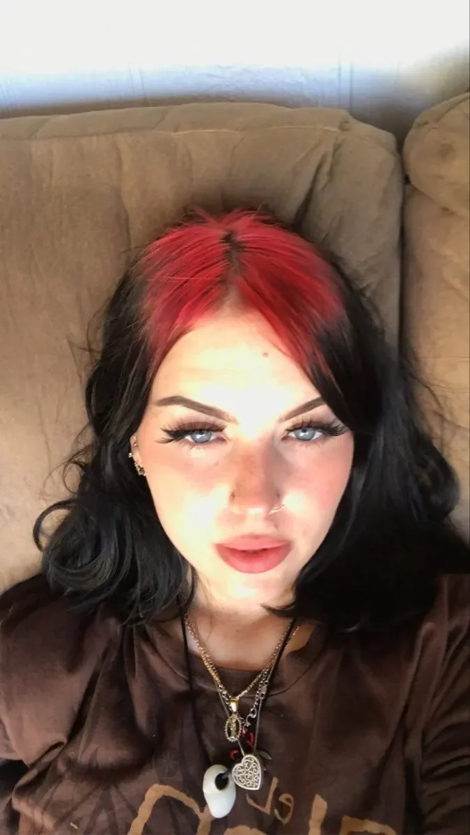 Woman with short soft black waves and two crimson-red root sections divided by the center part.