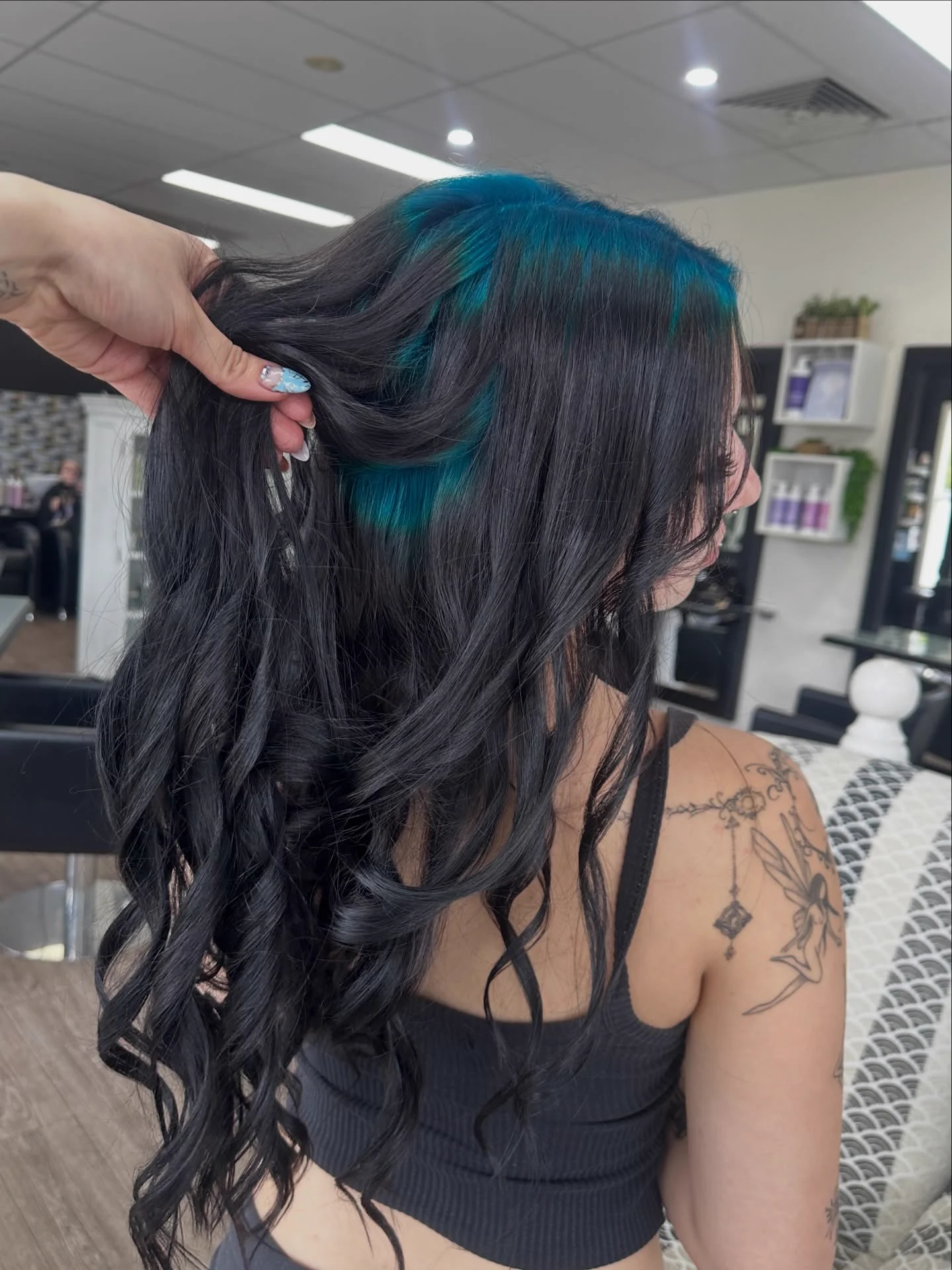 Long black wavy hair lifted at the side to reveal a broad teal root section beneath the top layer.