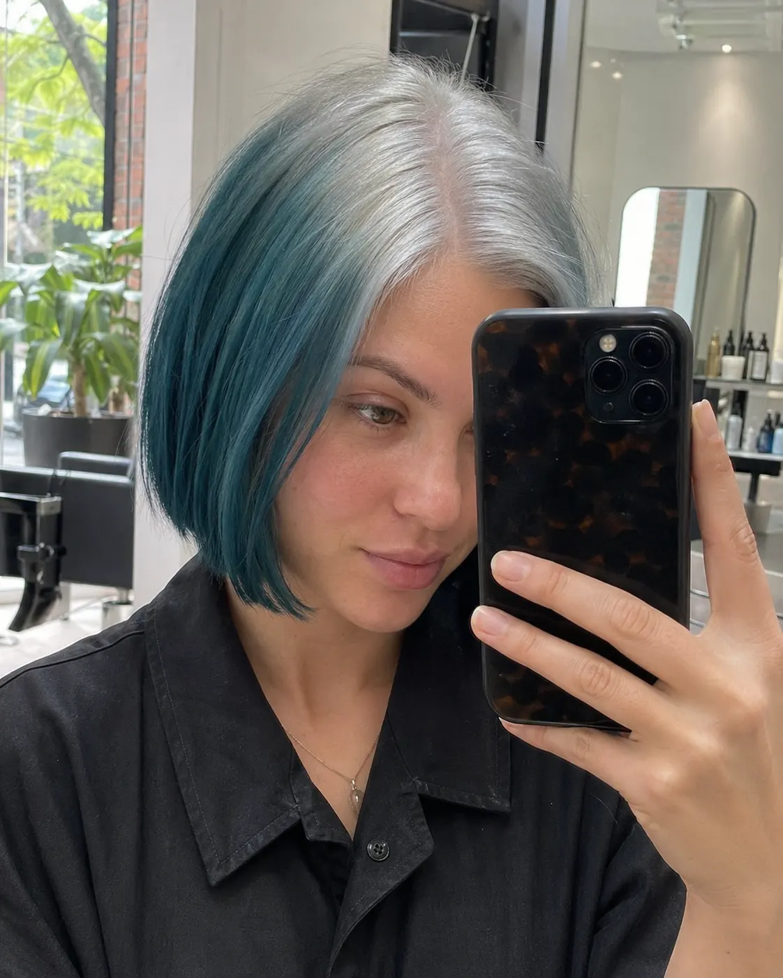 Woman with a short teal-blue sleek bob and silver-white color across the center-part and crown roots.
