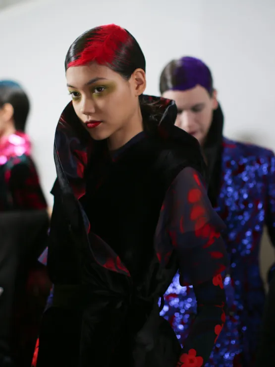 Backstage model at Dries Van Noten Fall 2020 with a red feather root illusion over slicked dark hair.