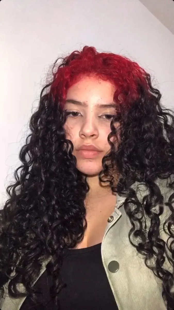 Woman with long black curls and crimson-red root color visible across the top and front crown.