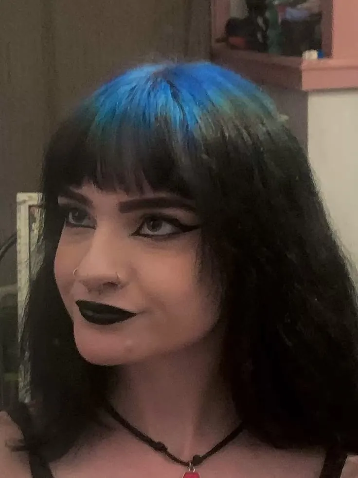 Long black straight hair with teal-blue ghost roots showing across the fringe and crown.